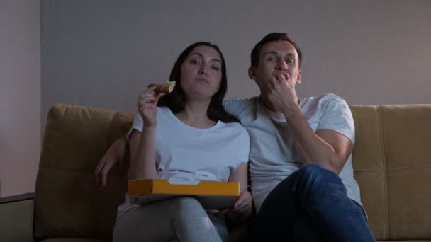 Мужчина и женщина едят пиццу и смотрят телевизор, сидя на диване — стоковое видео