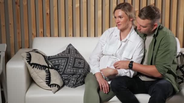 Hombre golpes embarazada señora vientre sentado en blanco sofá — Vídeos de Stock