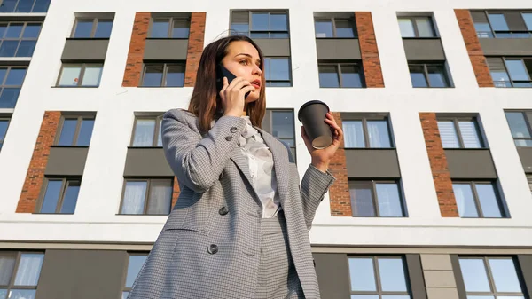 Femme en costume d'affaires boit du café dans une tasse en plastique et parle au téléphone sur le fond d'un bâtiment par une journée ensoleillée — Photo