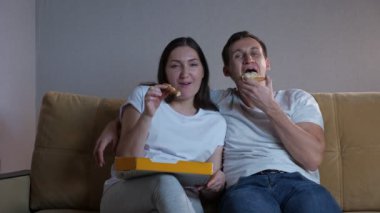 Kadın ve erkek pizza yer ve kanepede oturup televizyon izlerler.