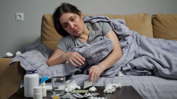 Больная женщина принимает лекарства и пьет воду, лежащую на диване — стоковое видео