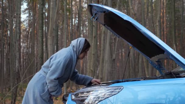 Žena se dívá pod kapotu rozbitého auta stojící lesní cesty — Stock video