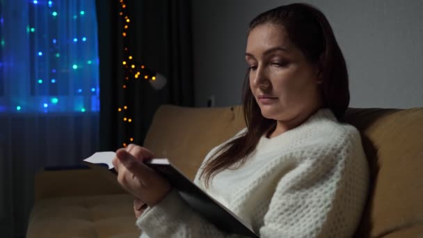 Ung kvinna läser bok sittandes på soffan i rummet på kvällen — Stockvideo