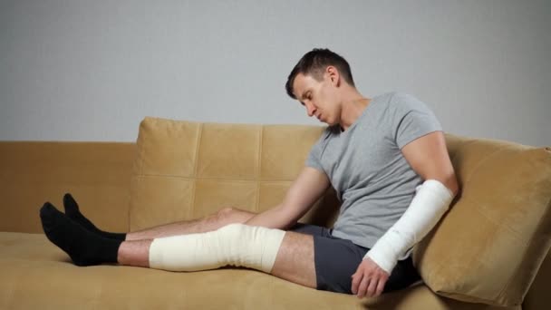 Man sits on sofa with injured knee and broken forearm — Stock Video