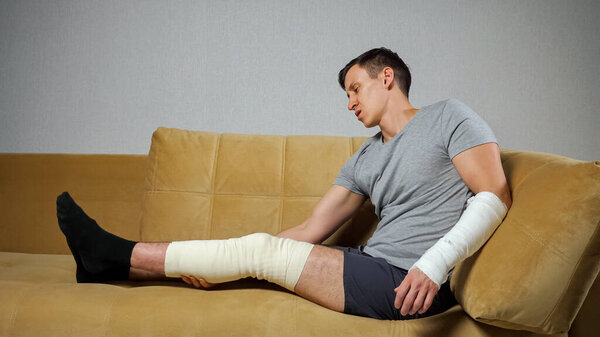 Мужчина сидит на диване с поврежденным коленом и сломанным предплечьем