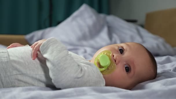 Piccolo bambino si trova sul letto morbido e si muove con manichino in bocca — Video Stock