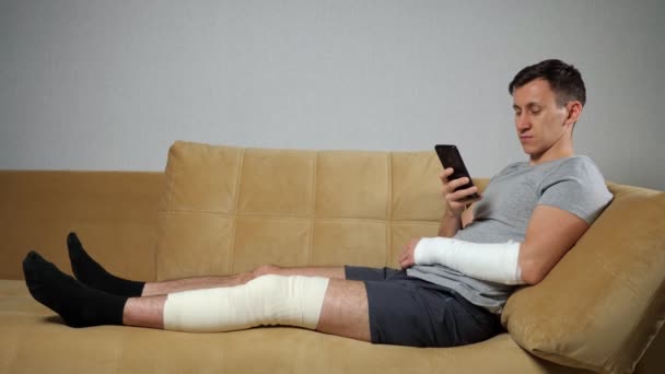 Hombre con la rodilla lesionada y el brazo roto se desplaza a través del teléfono — Vídeo de stock