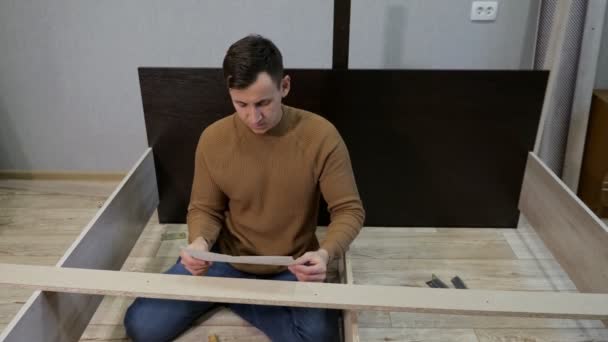 Young man in brown sweater assembles the bed according to the instructions — Stock Video