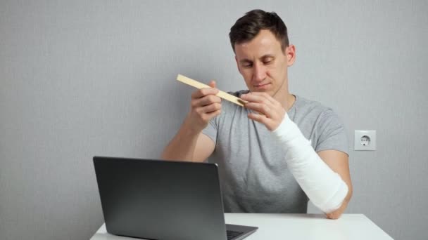 Young man scratches arm in gypsum band working on laptop — Stock Video