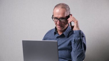 Yaşlı adam cep telefonuyla konuşuyor masada dizüstü bilgisayarla çalışıyor