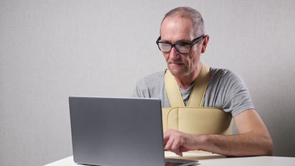 Hombre mayor con vendaje fijo trabaja en el portátil en el escritorio — Vídeos de Stock