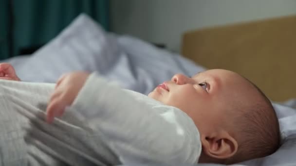 Baby boy lies on bed and moves blowing bubbles from saliva — Stock Video
