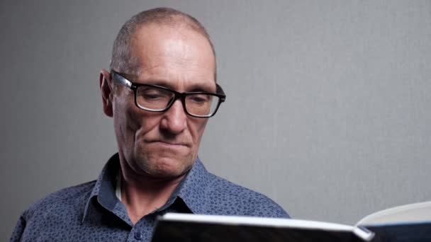 Hombre mayor en gafas elegantes lee libro sobre fondo gris — Vídeo de stock