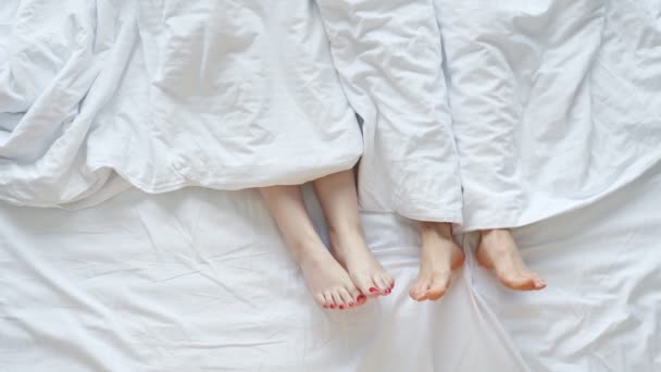 Jóvenes y mujeres yacen en la cama con las piernas fuera de la sábana — Vídeo de stock