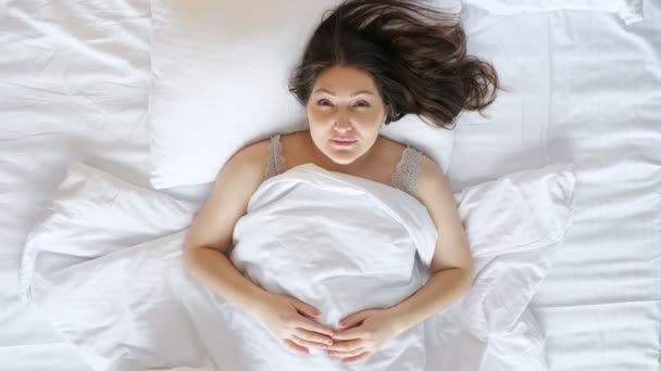 Mujer joven sonríe extensamente acostada en la cama con sábanas blancas — Vídeo de stock