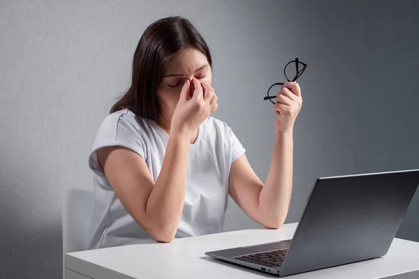 Young woman massaging the bridge of nose, holding glasses in other hand, sitting in front of a laptop — Stockfoto