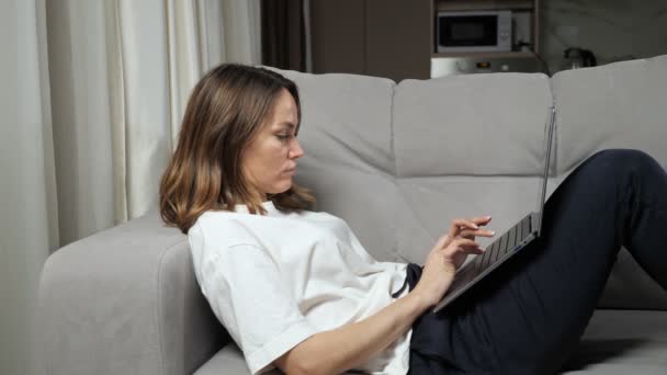 Freelancer mujer trabaja en línea a través de ordenador portátil tumbado en sofá gris — Vídeos de Stock