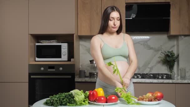 Zwangere vrouw peelt en eet selderij strelen blote buik — Stockvideo