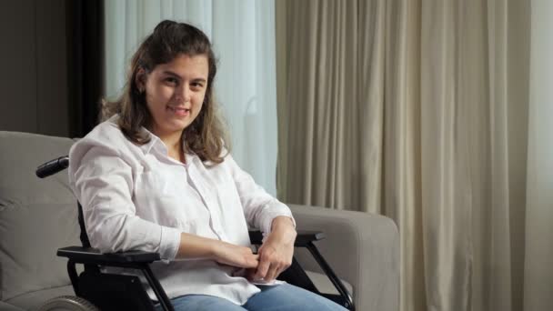 Mujer discapacitada sonríe sentada en silla de ruedas por sofá en casa — Vídeos de Stock