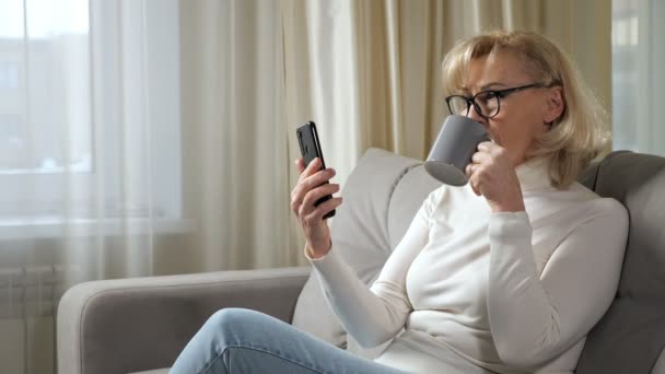 Mujer madura sorbe café y lee noticias en línea por teléfono — Vídeos de Stock