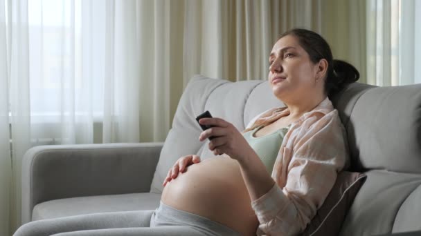 Femme enceinte ennuyée commute les chaînes de télévision caressant ventre — Video