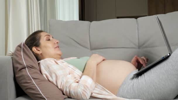 Беременная мать делает онлайн покупки для ребенка, лежащего на диване — стоковое видео