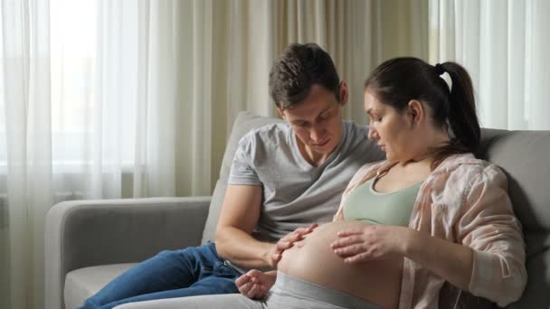 Муж ласкает и целует беременный желудок жены на диване — стоковое видео