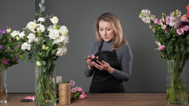 Mujer florista rubia utiliza teléfono móvil en taller de floristería — Vídeos de Stock