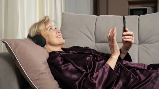 Ηλικιωμένη γυναίκα διαβάζει ειδήσεις ακούγοντας μουσική μέσω ακουστικών — Αρχείο Βίντεο