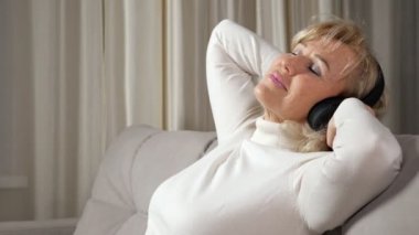 Orta yaşlı sarışın kadın kulaklıkla müzik dinliyor.