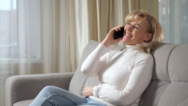 Mujer de cabello rubio habla a través de teléfono inteligente y sonríe en el sofá — Vídeos de Stock