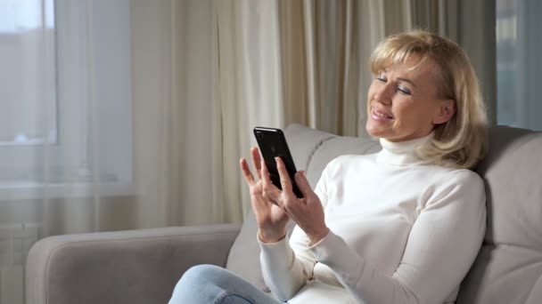 Mujer anciana sonríe felizmente después de leer buenas noticias en el sofá — Vídeos de Stock