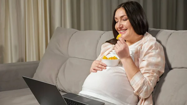 Brunette woman enjoys junk food watching show via laptop — Stock Photo, Image