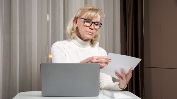 Женщина среднего возраста в очках считает счета за коммунальные услуги за столом — стоковое видео