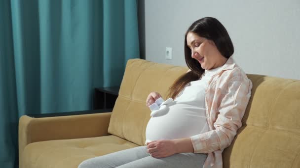Pregnant woman holding white booties in hand stroking belly while sitting on the couch — Stock Video