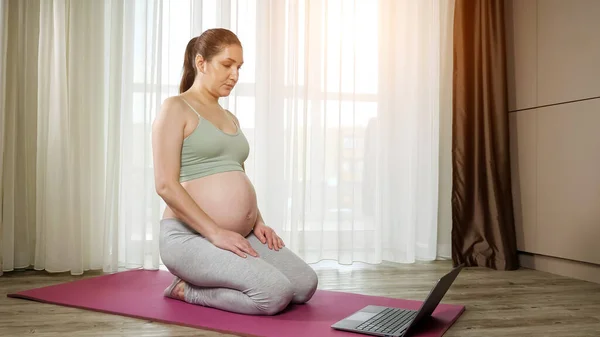 Pregnant woman warms up watching tutorial via laptop on mat — Stock Photo, Image
