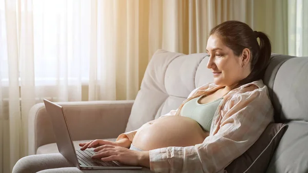 임신 한 여성 프리랜서는 소파에서 노트북을 사용하여 집에서 일한다 — 스톡 사진