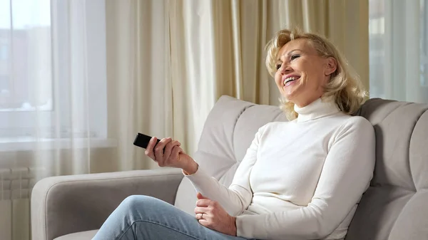 Elderly blonde woman laughs and enjoys watching TV series — Stock Photo, Image
