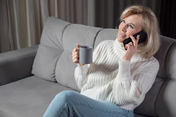 Mulher de cabelos loiros fala via smartphone e sorri no sofá — Fotografia de Stock