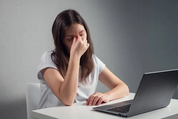 Mujer joven masajeando el puente de la nariz y los ojos mientras está sentada frente a una computadora portátil Imágenes de stock libres de derechos