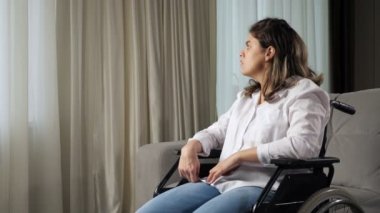 Merkezi sinir sisteminde organik hasarı olan üzgün bir kadın tekerlekli sandalyede oturmuş evdeki oturma odasındaki perdenin arkasına bakıyor..