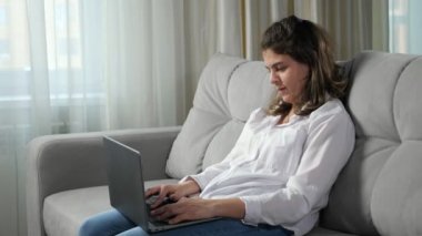 Serebral felçli genç bir kadın bilgisayarla uzaktan çalışıyor evdeki pencere pencere kenarındaki kanepede oturan cep telefonuyla mesaj yazmaya çalışıyor..