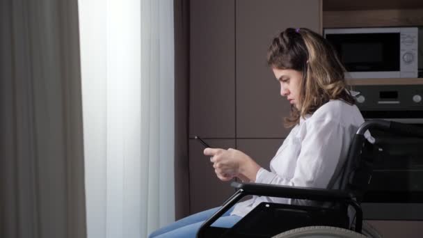 白いシャツを着た障害のある女性は カーテンの後ろの窓の近くの車椅子に座っているスマートフォンでメッセージを入力しようとしますホームサイドビュー — ストック動画
