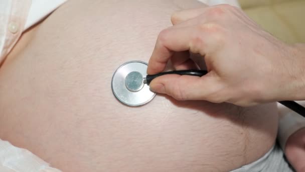 Mão de um homem irreconhecível dirige um estetoscópio sobre a barriga grande de uma mulher grávida — Vídeo de Stock