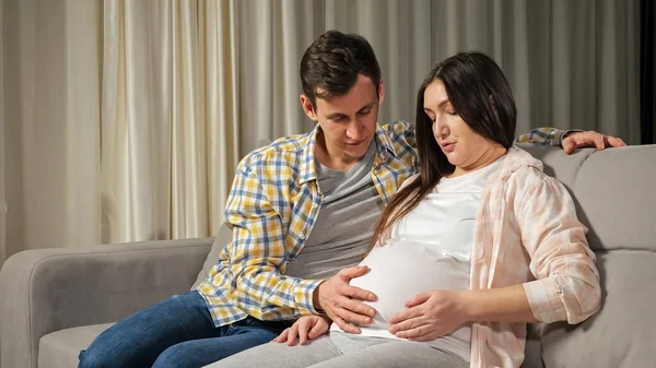 Il marito accarezza lo stomaco incinta di moglie seduta sul divano — Foto Stock