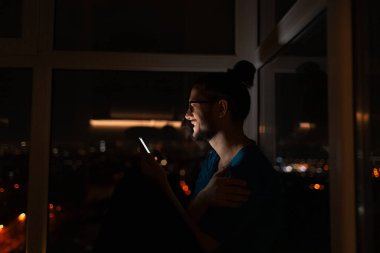 Pencerenin yanında elinde akıllı telefonu olan mutlu bir gencin gece portresi..