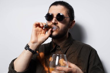 Beyaz arka planda puro ve viski içen özgüvenli bir adamın stüdyo portresi..