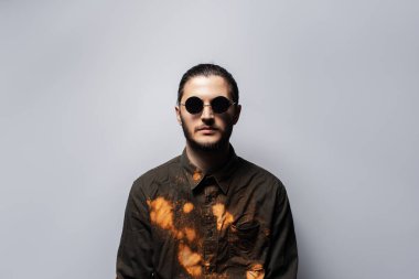 Gri arka planda yuvarlak güneş gözlüğü takan kendinden emin genç bir adamın portresi..