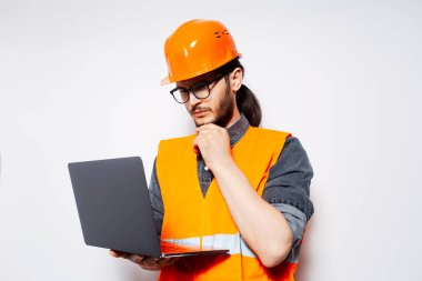 Genç düşünceli mühendis dizüstü bilgisayara bakıyor, turuncu güvenlik ekipmanları giyiyor..