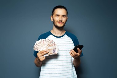 Portrait of young man holding money, Moldovan banknotes and smartphone, on blue.